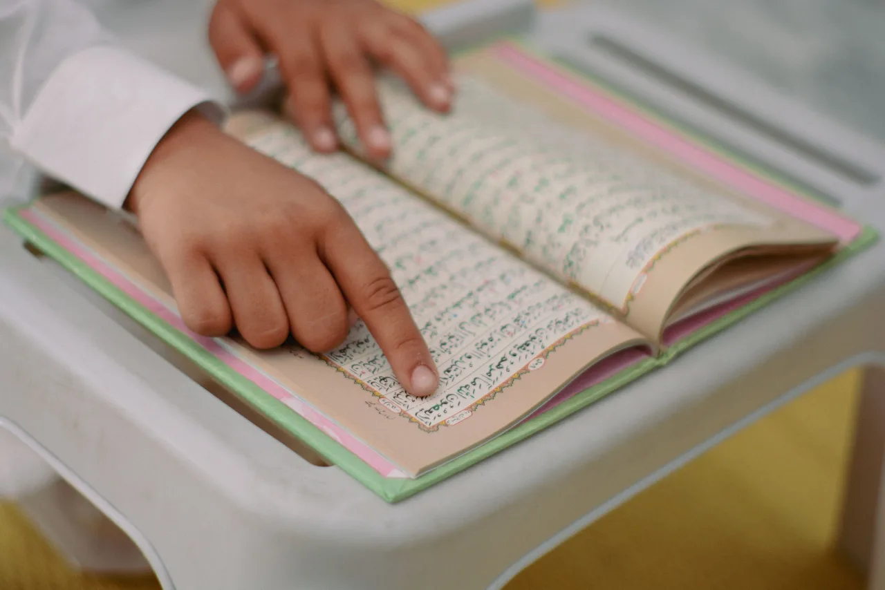 A young child reading Arabic from an open, colourful Quran at home, following the words with their finger
